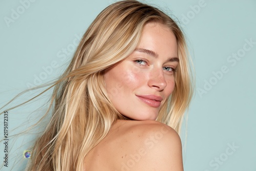 Close-up portrait of a young fair-haired woman with freckles smiling softly as she looks thoughtfully away from camera, long blonde hair gently moving as if caught by breeze