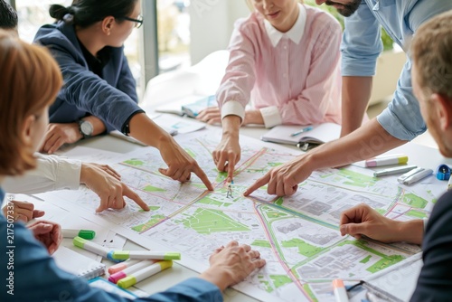 Group of diverse professionals gathered around a large urban development map on a white table pointing at transportation routes green zones and building districts with colorful removable markers