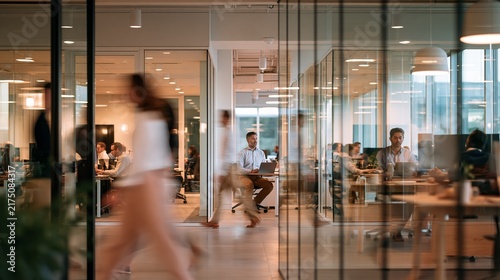 Several employees moving through glass office partition with intentional motion blur while one man remains sharply focused at laptop workstation under mixed daylight and soft overhead lighting in
