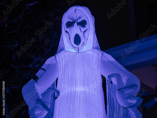 A glowing ghost inflatable with a haunting face stands outside during Halloween evening. Blue and purple lighting adds a dramatic spooky atmosphere.