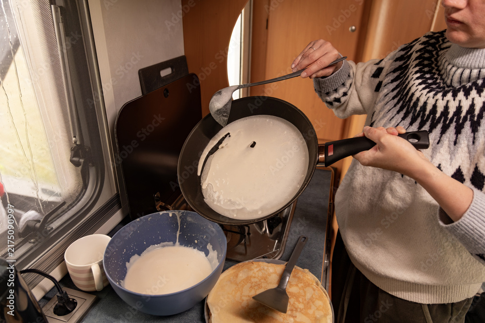 Fototapeta premium Woman cooking pancakes in a caravan kitchen, holding a frying pan with batter, bowls of pancake mix and utensils visible on the countertop