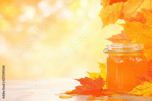 Glass honey jar with vibrant autumn maple leaves on golden bokeh background with copy space.