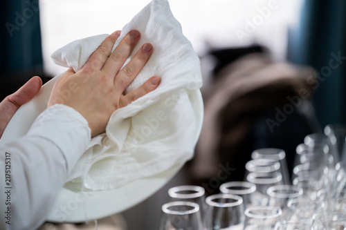 A close-up of a person's hands using a white cloth to polish and dry a white ceramic plate, with rows of clean wine glasses standing in the blurred background.