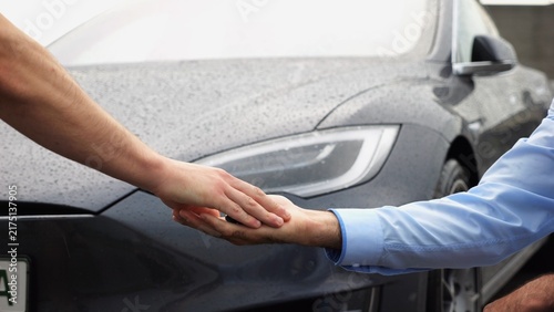 Male arm giving key of car to buyer of electric automobile at rainy day. Handshake between salesman and new owner of modern auto after successful deal. Concept of buy or rent car. Slow motion