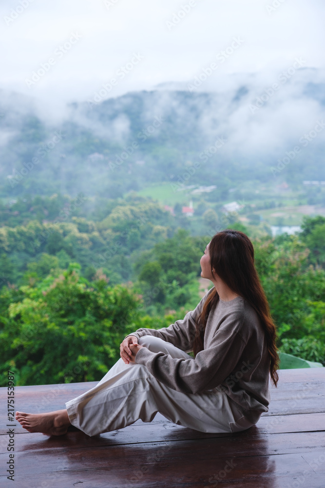 Naklejka premium Vertical image of a woman sitting on wooden balcony with a beautiful mountain views on cloudy morning