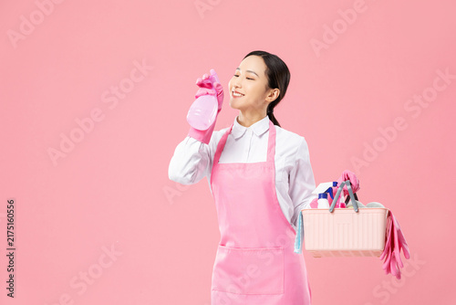 Smiling housekeeper with spray bottle and cleaning supplies on pink background