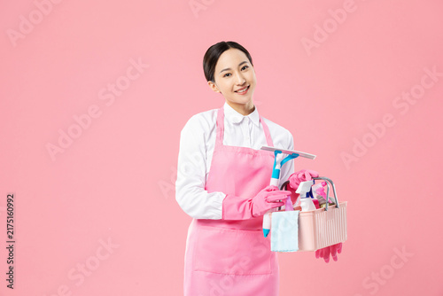 Smiling housekeeper holding cleaning supplies on pink background