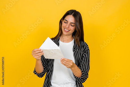 Woman smiling with closed eyes, holding an open envelope with a letter or important message inside, standing against a bright yellow background, conveying happiness and positive news