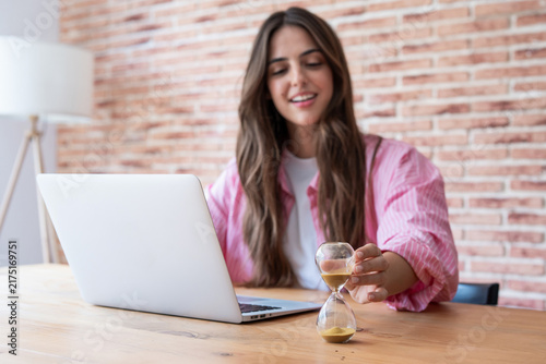 A young woman working at a laptop in a cozy home office turns an hourglass to track time, suggesting productivity, deadlines, time management, focus and remote work balance in a relaxed setting.