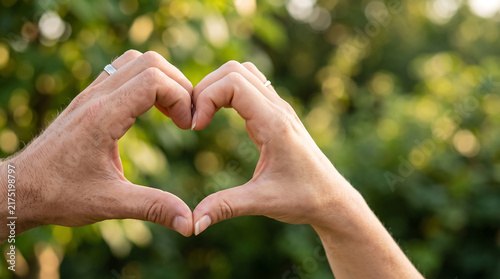 Close up of two hands creating a heart shape against a blurred green outdoor background, representing love, romance, affection, connection, partnership and togetherness.
