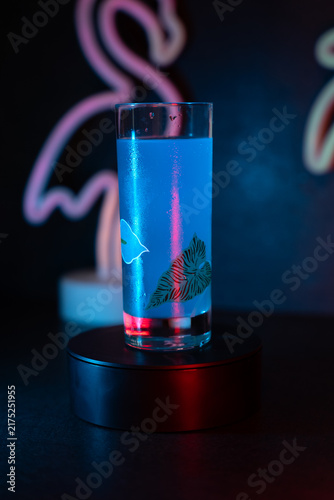 Blue liquid glows in a tall glass. Neon pink flamingo shines in the background. Red and blue lights create a moody atmosphere. Glass sits on a black rotating base. Ideal for nightlife, party