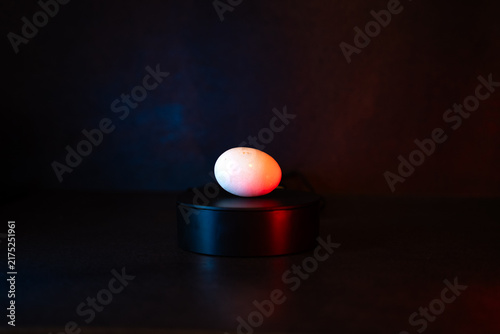 Single egg rests atop sleek black pedestal. Soft red glow wraps its smooth, rounded shell. Dark background highlights egg's subtle warmth. Minimalist setup suggests mystery or stillness