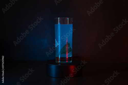 Glass holds glowing liquid. Red light casts soft shadows. Blue glow illuminates mysterious shape. Base rotates slowly, creating movement. Dark background enhances visual focus