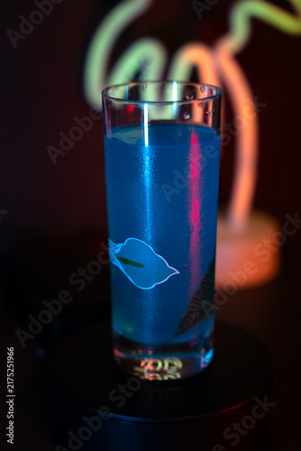 Blue drink glows under neon lights. Lips design floats in the liquid. Dark background enhances drink's glow. Neon sign adds moody ambiance. Perfect for nightlife or cocktail scenes