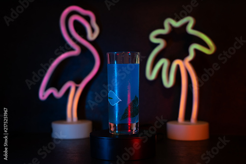Neon flamingo glows softly beside palm tree. Blue drink sits cool on dark stand. Vibrant lights cast colorful shadows on dark surface. Perfect for tropical, nightlife, or modern bar scenes