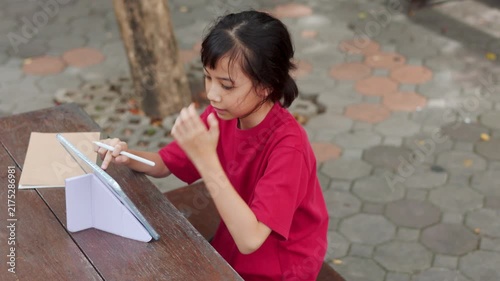 Asian kid using stylus pen on a digital tablet for e-learning at outdoor school table. Selective focus shows the young student engaging with technology for modern education and digital study concepts.