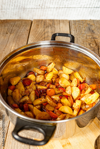 Sauteed potatoes with sliced sausage, carrots, and spices mixing in a stainless steel cooking pot, rustic wooden background, meal preparation step.