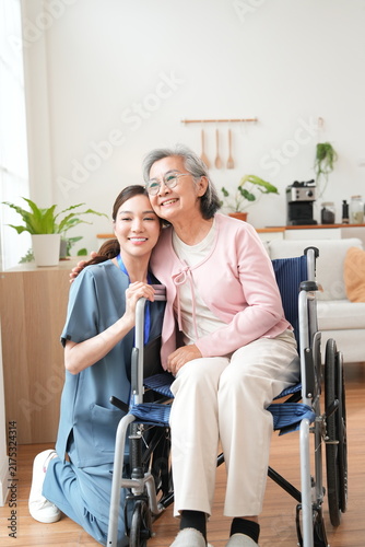 An elderly female patient with Alzheimer's disease is in a wheelchair, with an Asian nurse sitting beside her, holding her hand, offering encouragement, and pushing the wheelchair, caring for the elde