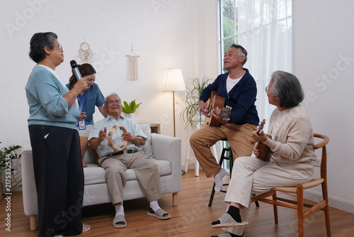 A group of elderly people are preparing for an activity: a senior woman holds a microphone and sings, and her friend plays guitar, enjoying music therapy at the nursing home.