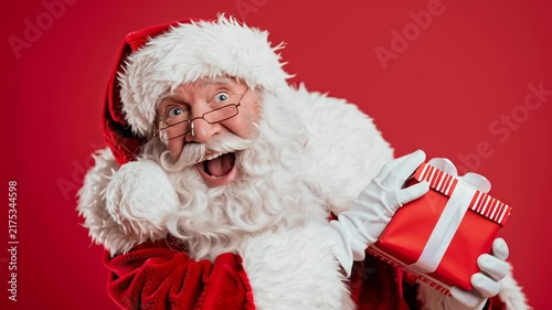 Santa Claus smiles broadly, holding a gift. His eyes gleam with joy as he points cheerfully. Red background highlights his festive spirit. Perfect for holiday campaigns or gift-related content