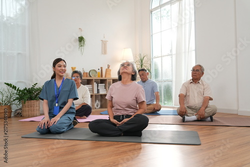 A group of elderly men and women are exercising and practicing yoga, with an Asian female nurse serving as a turner providing physical support and care in a class at the nursing home.