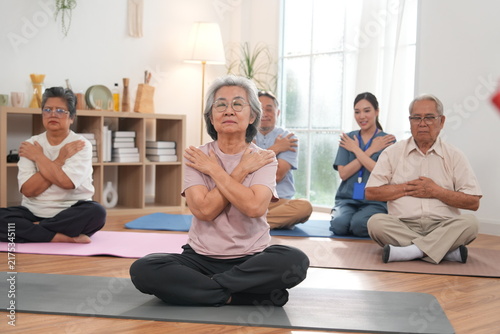 A group of elderly men and women are exercising and practicing yoga, with an Asian female nurse serving as a turner providing physical support and care in a class at the nursing home.