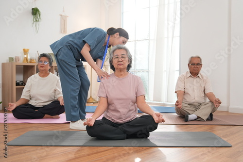 A group of elderly men and women are exercising and practicing yoga, with an Asian female nurse serving as a turner providing physical support and care in a class at the nursing home.