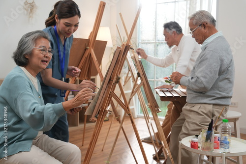 Elderly men and women in the nursing home engage in art therapy activities for relaxation and rest, and an Asian female nurse uses paintbrushes to create paintings, demonstrating her skills.