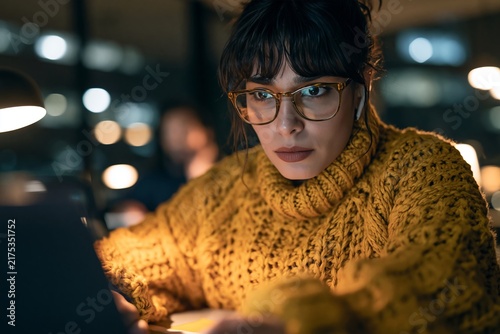 Focused woman wearing glasses and a yellow sweater working late at night in an office setting