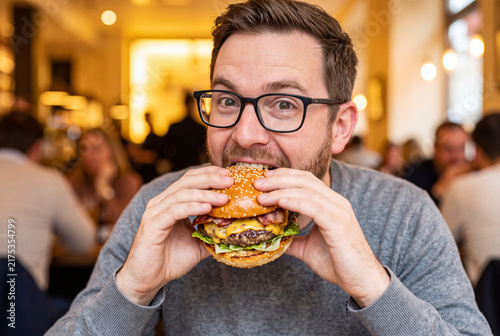 Excited Young Man Taking a Large Bite of Juicy Cheeseburger in Restaurant
