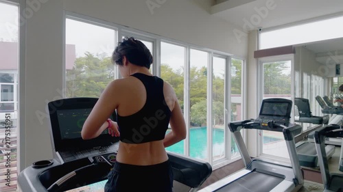 Rear view of an active Asian woman in her forties wearing a black sports bra while running on a treadmill. She exercises in a modern gym, looking out large windows during her daily cardio workout.