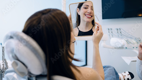 Patient smiling in a dental chair, looking at her new veneers in a mirror at the modern dental clinic