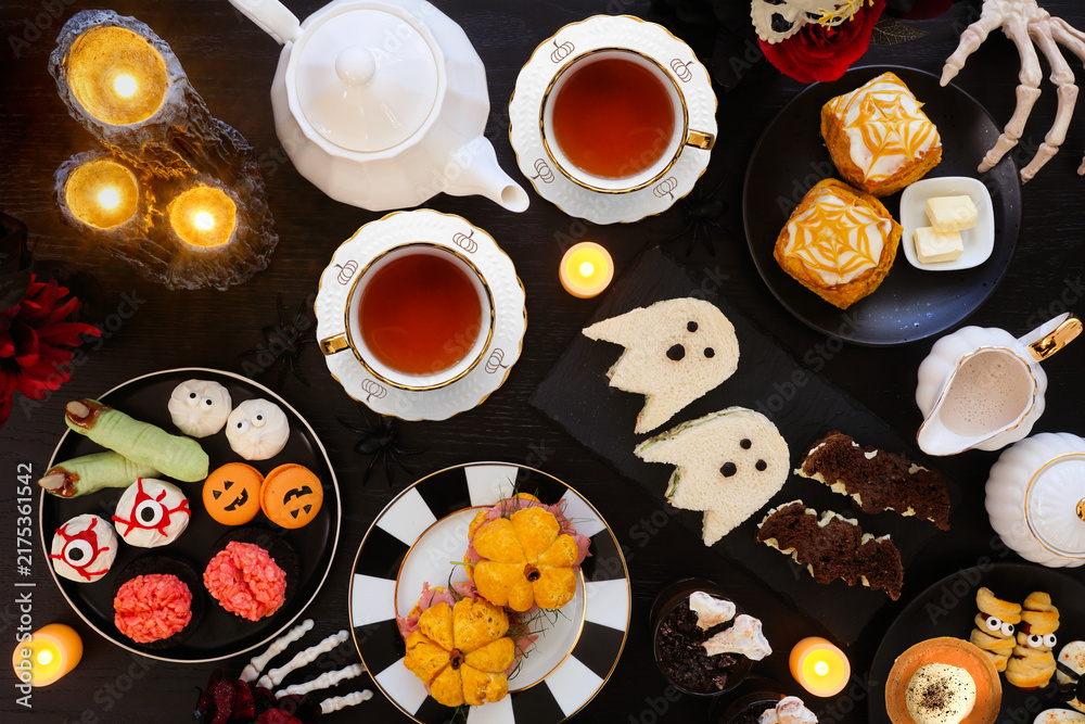 Obraz premium Halloween theme afternoon tea table scene against a black wood background. Spooky savory finger foods, sandwiches, pumpkin scones, and scary desserts. Top down view.