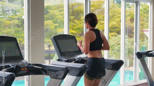 Wide shot of an active Asian woman in her forties running on a treadmill in a bright modern gym. She wears a black sports bra, enjoying daily cardio exercise by large windows with natural daylight.