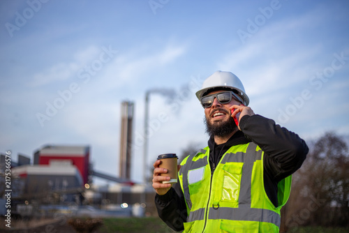 Engineer working in industrial area, smiling while talking on phone and holding a coffee cup, with power plant in background