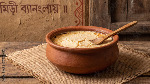 Traditional Bengali mishti doi (sweet yogurt) served in a terracotta pot with a wooden spoon, textured backdrop