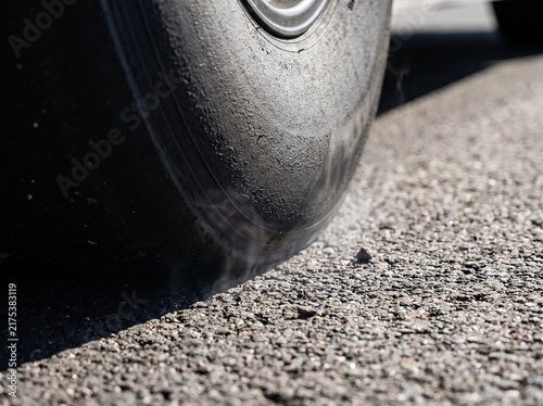 Heavy black airplane tire emits visible smoke while landing on rough asphalt runway under bright natural daylight conditions during arrival