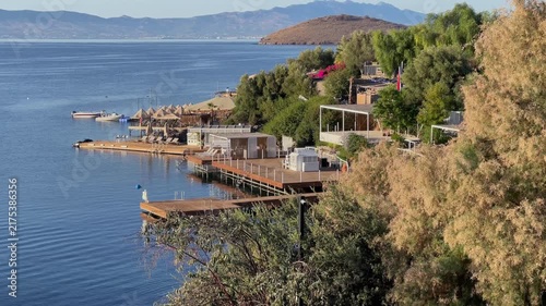 A sunny elevated view of a coastal resort featuring wooden piers, sun loungers, and beach cabanas along a tranquil blue bay with distant mountains.