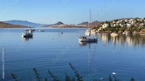 A calm blue bay filled with anchored sailboats and yachts, set against a backdrop of a white coastal village, rolling hills, and a clear blue sky.