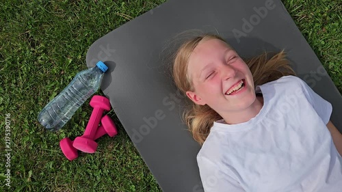 Girl in white t‑shirt laughing, lying on grey mat outdoors. Pink dumbbells and plastic water bottle on grass. Bright daylight, joyful mood, carefree moment