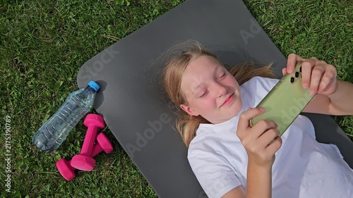Girl in white t‑shirt smiling, holding smartphone, lying on grey mat outdoors. Pink dumbbells and plastic water bottle on grass. Natural daylight, relaxed atmosphere, fitness break.