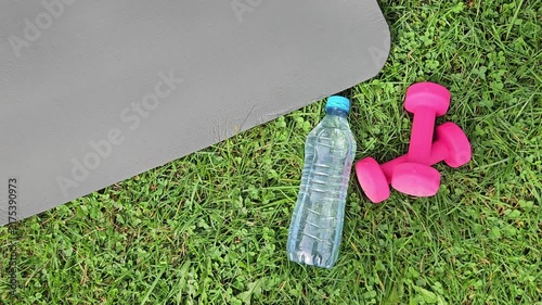 Close‑up outdoor shot showing a dark grey fitness mat partially in frame on the left, its smooth textured surface angled diagonally. On green grass in the foreground — a transparent plastic water bott