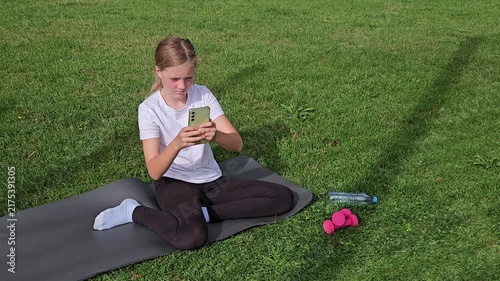 Girl in white t‑shirt and black leggings sitting on grey yoga mat on grass, looking at smartphone. Pink dumbbells and plastic water bottle on grass. Bright daylight, relaxed outdoor atmosphere