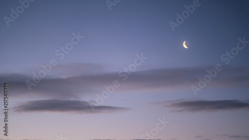 A thin, bright crescent moon is visible in a soft, gradient sky transitioning from pale blue to dusky purple, with delicate, horizontal streaks of grey clouds adding texture and depth to the serene