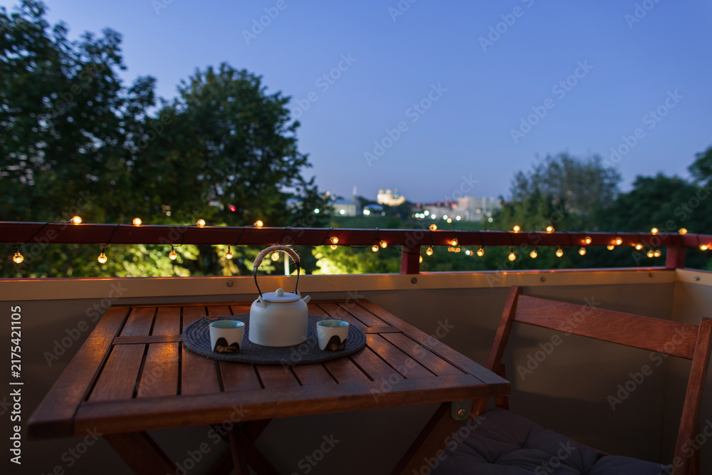 Obraz premium Cozy balcony setup at twilight with warm string lights on the railing, a wooden table with ceramic teapot and cups, offering a serene city view for a relaxing evening atmosphere.
