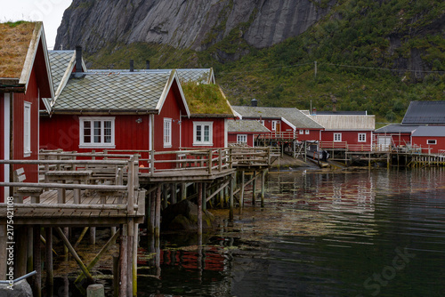 Traditionelle rote Rorbuer Hütten auf Pfählen mit Holzstegen und Grasdächern im Fischerdorf Reine vor steiler Felswand, Reine, Lofoten, Norwegen, Europa