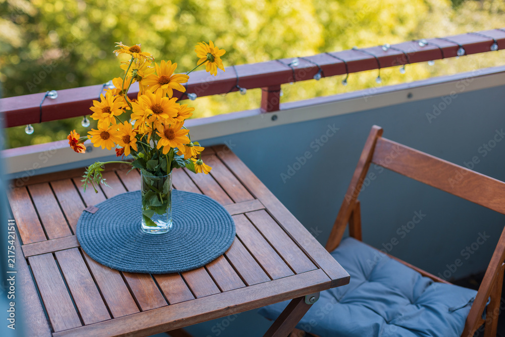 Obraz premium Bright yellow flowers in a glass vase on a wooden balcony table with a cozy chair, placemat, string lights, and warm sunlight creating a charming outdoor setting. Autumn day