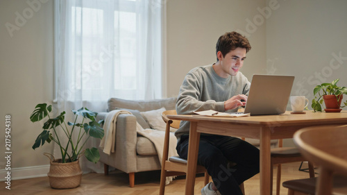 Young man focused on his laptop at a wooden dining table in a cozy living room, with a sofa, plants, and soft curtains, depicting a comfortable remote work or study environment