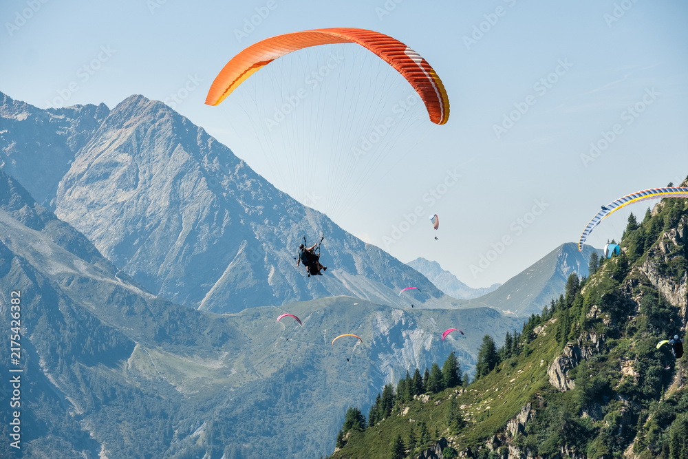 Obraz premium Paraglider flying with bright orange wing high above mountain valley in Chamonix French Alps during scenic summer day with extreme tandem flight adventure travel experience over alpine landscape