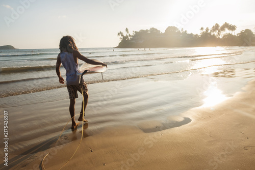 Black long haired man holding white surfboard walking on wet sand towards ocean waves at beautiful Dewata beach in Sri Lanka during warm golden hour sunset enjoying active summer surfing holiday